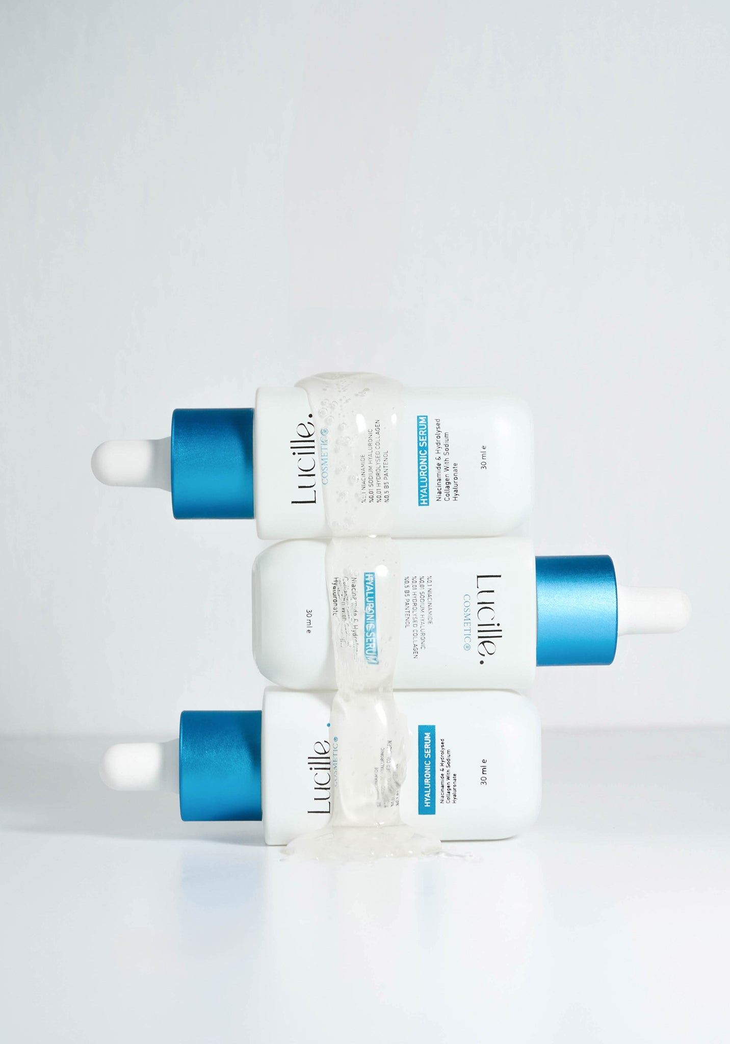 Four white Lucille skincare dropper bottles stacked in a 2x2 grid, each with a metallic blue cap, on a clean white background with a translucent clear wrap around the center.