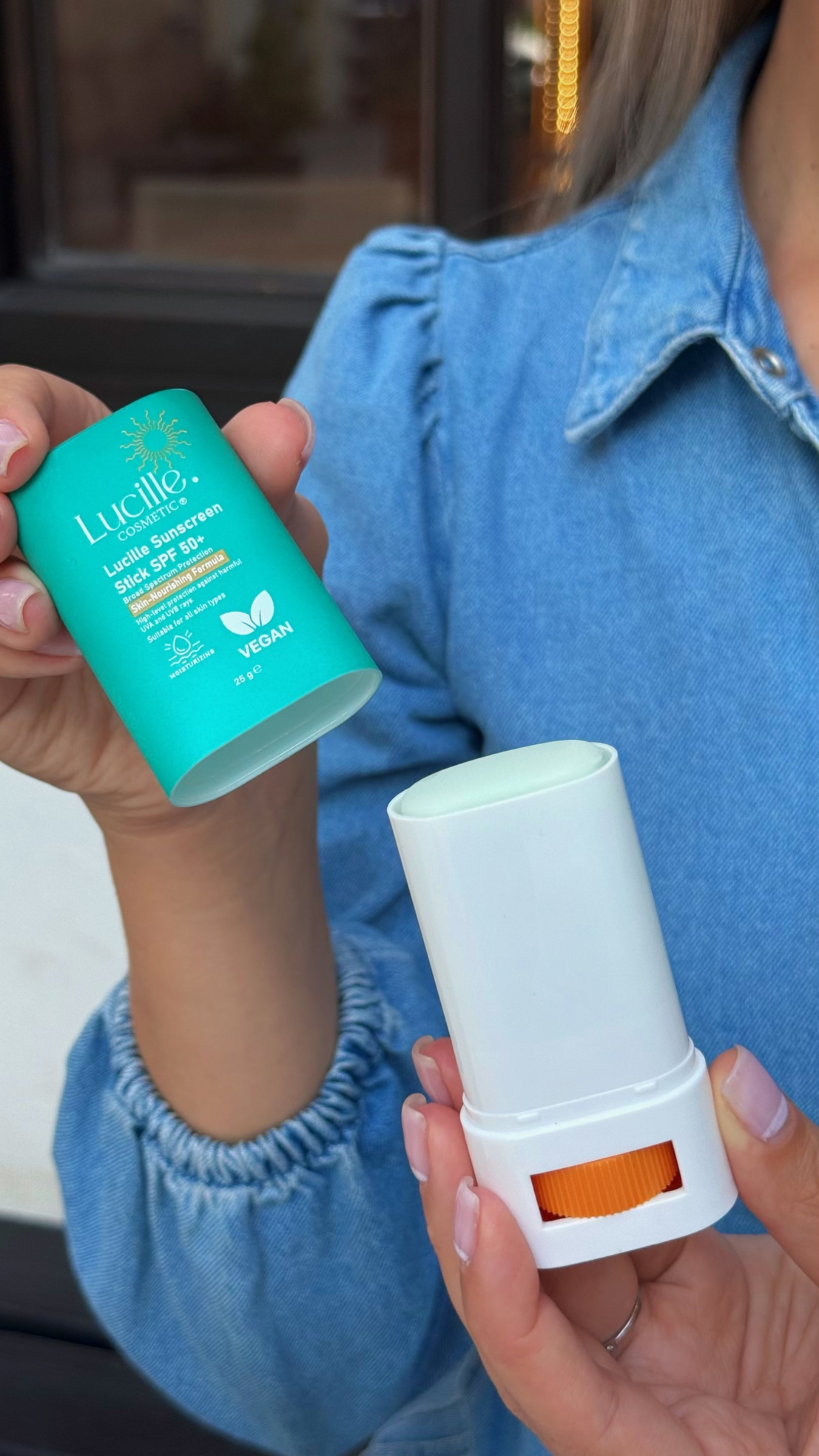 A person in a blue denim shirt holds a turquoise Lucille Light Blocking SPF 15 stick in one hand and a white reusable sunscreen stick container with an orange window in the other.