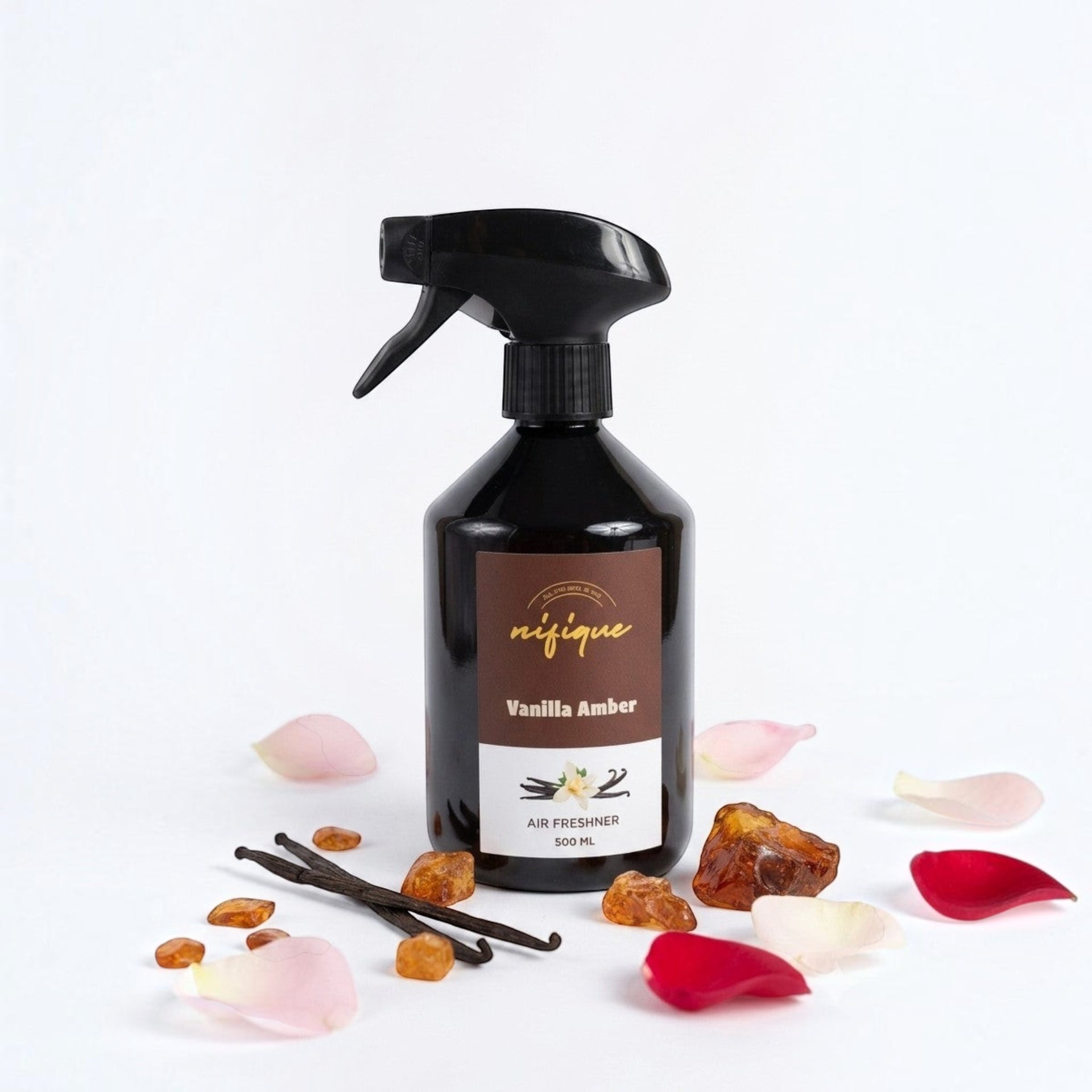 A 500 ml black spray bottle of 'nique' Vanilla Amber air freshener, styled with vanilla beans, amber crystals, and pink and red rose petals on a clean white background.